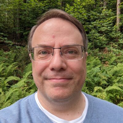 Headshot photo of me wearing glasses and a blue sweater with green forest behind me.